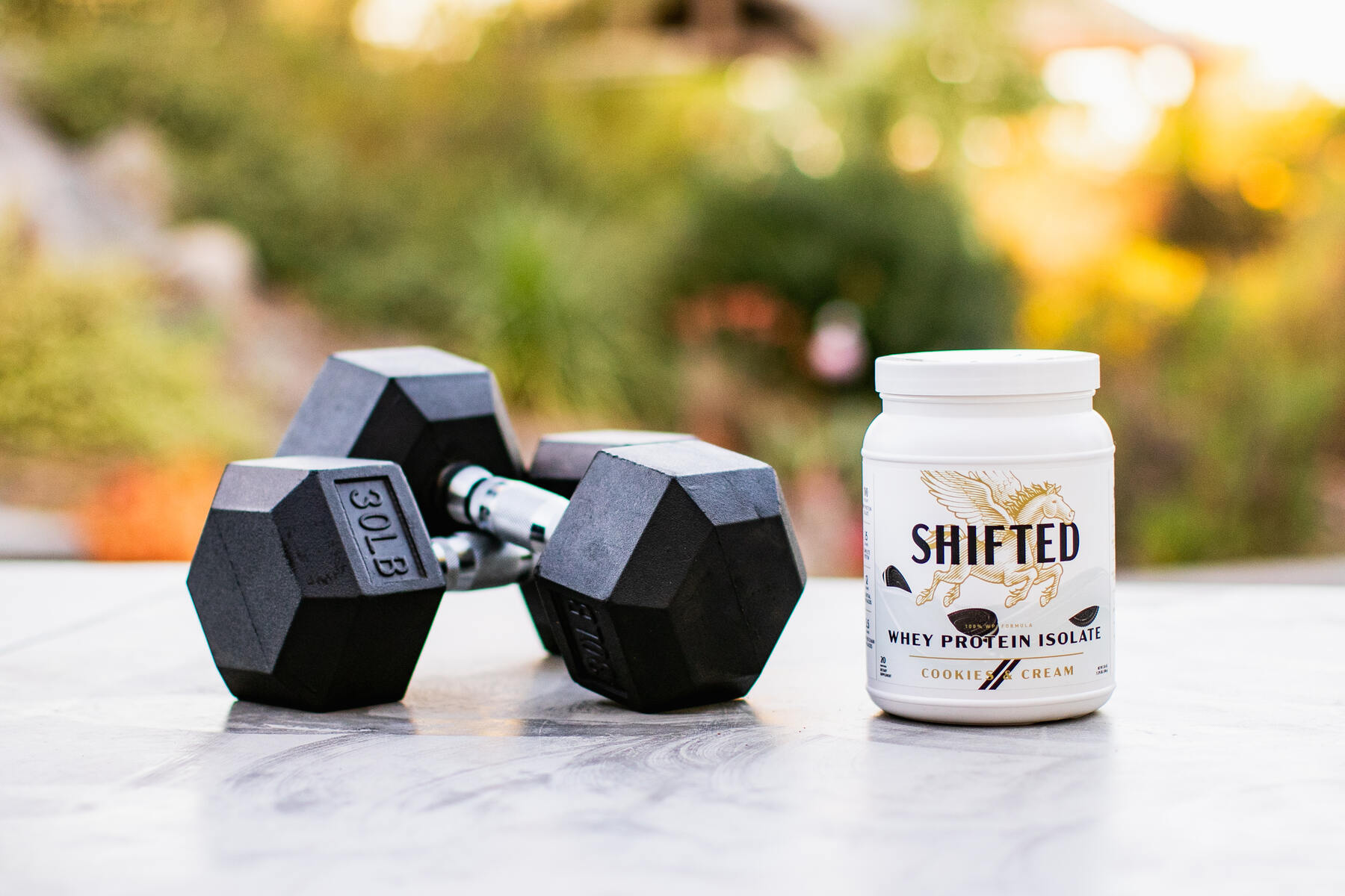Shifted whey protein isolate next to 30lb dumbbells on a gray concrete table outside