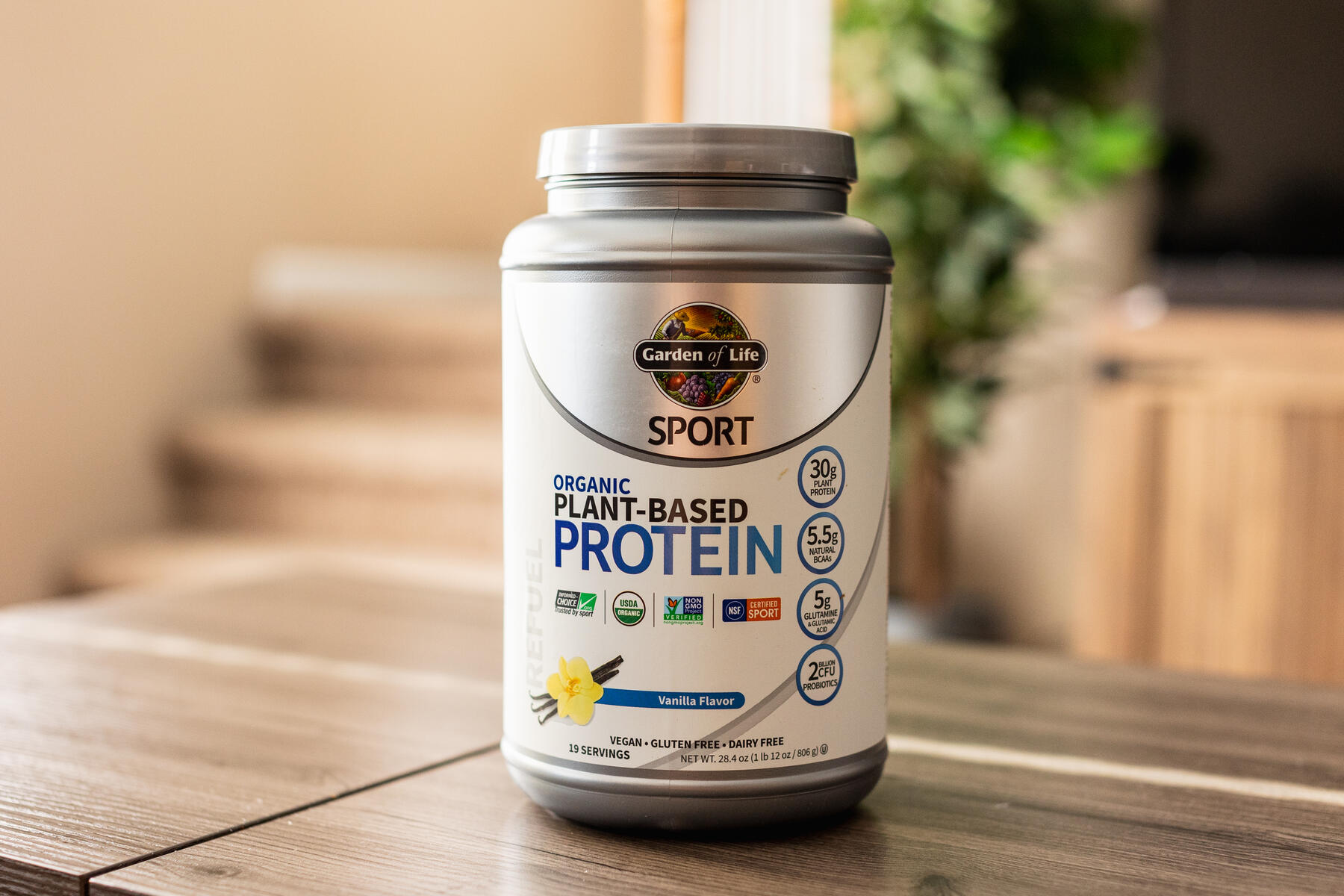 A silver container of Garden of Life Sport Organic Plant-Based Protein powder in vanilla flavor, placed on a wooden surface with greenery in the background