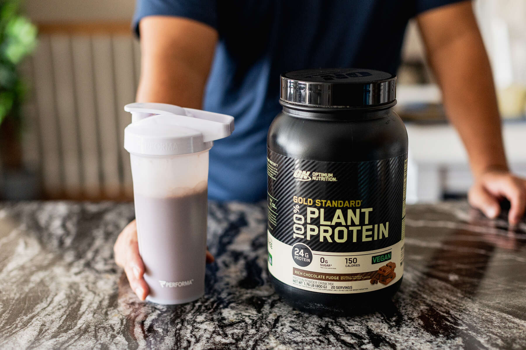 A person in a blue shirt holding a white protein shaker with a black container of Optimum Nutrition Gold Standard Plant Protein on a marbled countertop