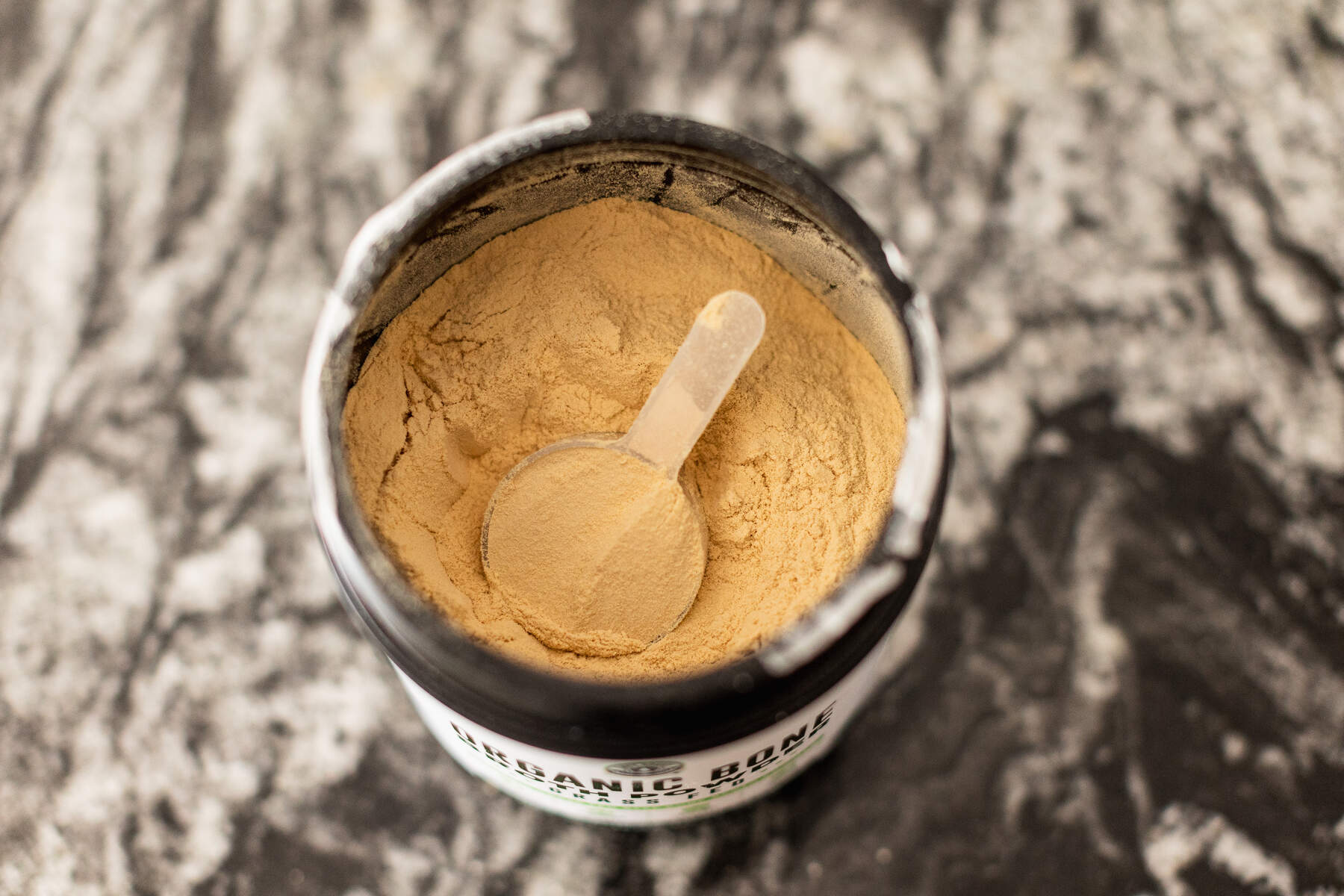 Open jar of protein powder with a clear scoop inside of it, placed on a granite countertop