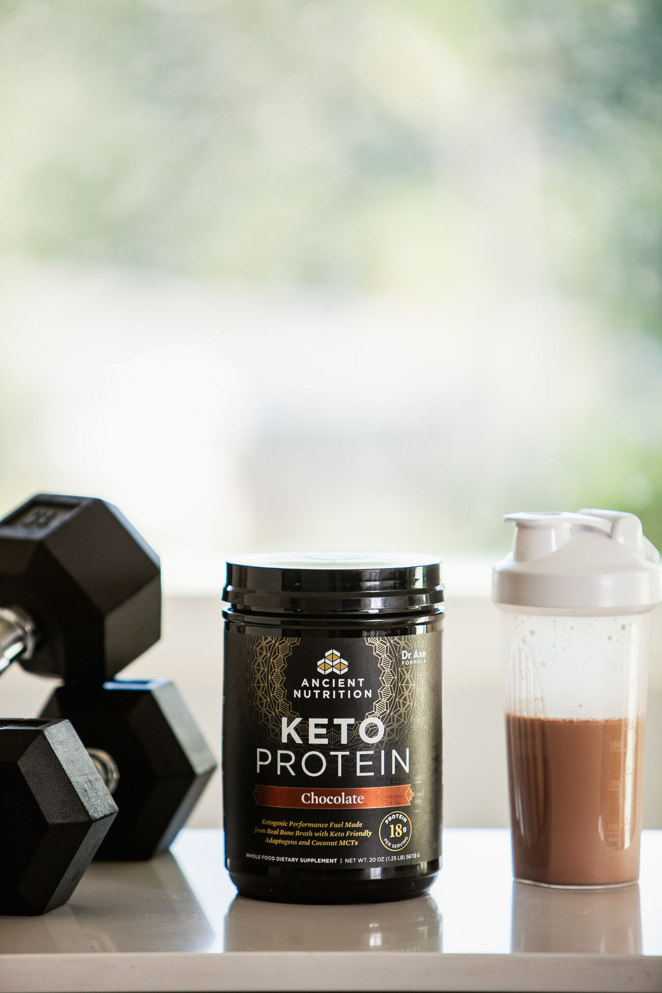 Chocolate keto protein power next to a protein shake and a pair of dumbbells on a white table