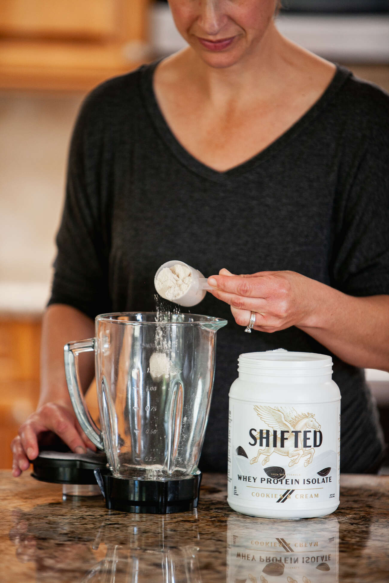 A woman scooping whey protein powder into a blender on a kitchen counter