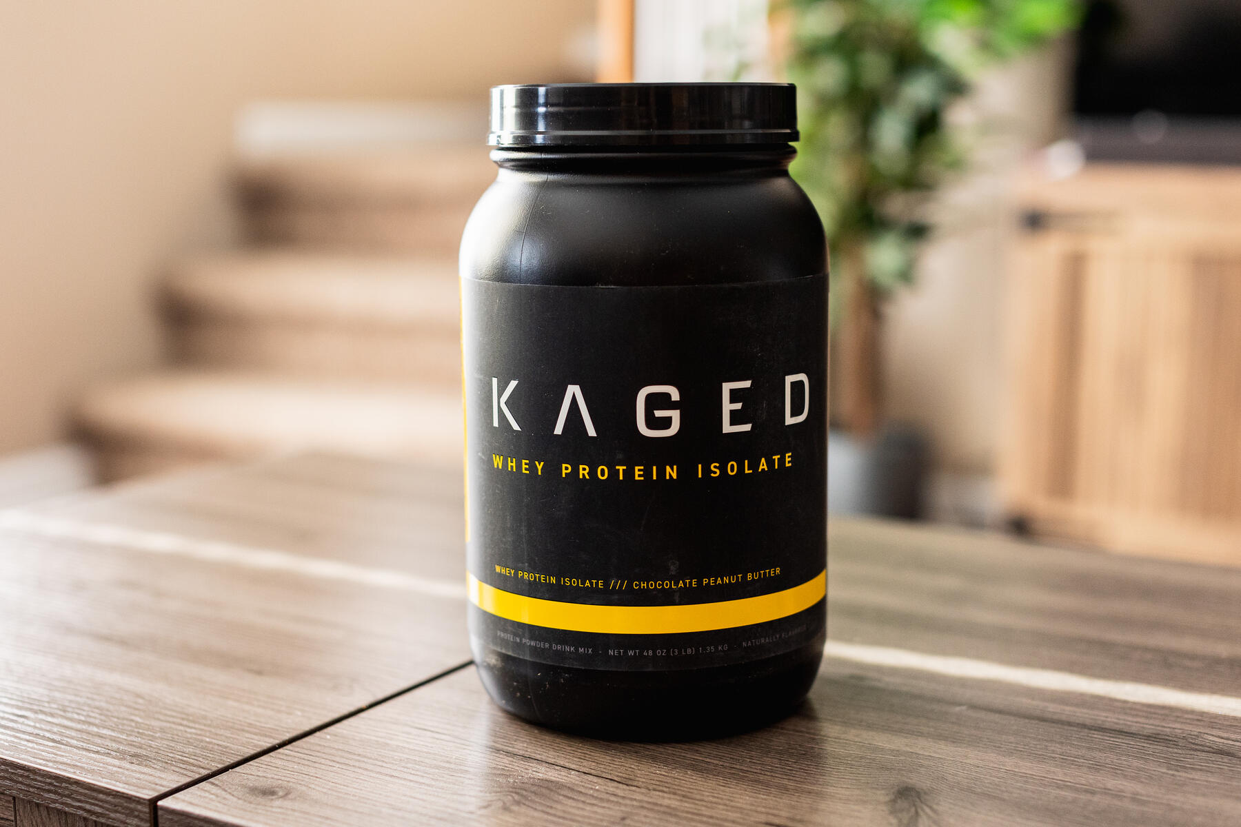 A jar of whey protein isolate on a wooden table with a plant in the background