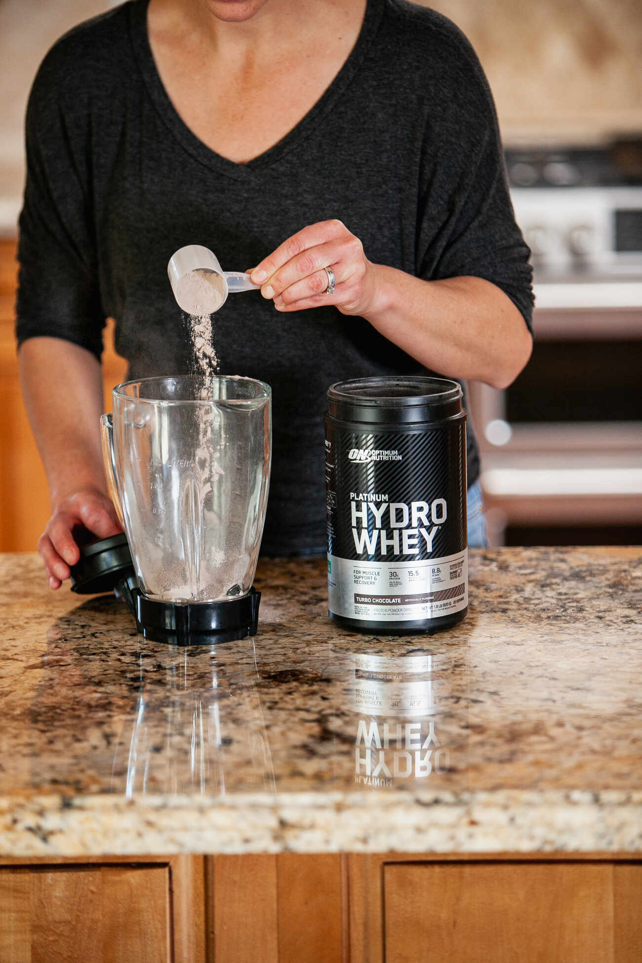 A woman pouring chocolate-flavored whey protein powder from a scoop into a blender, with the container in view