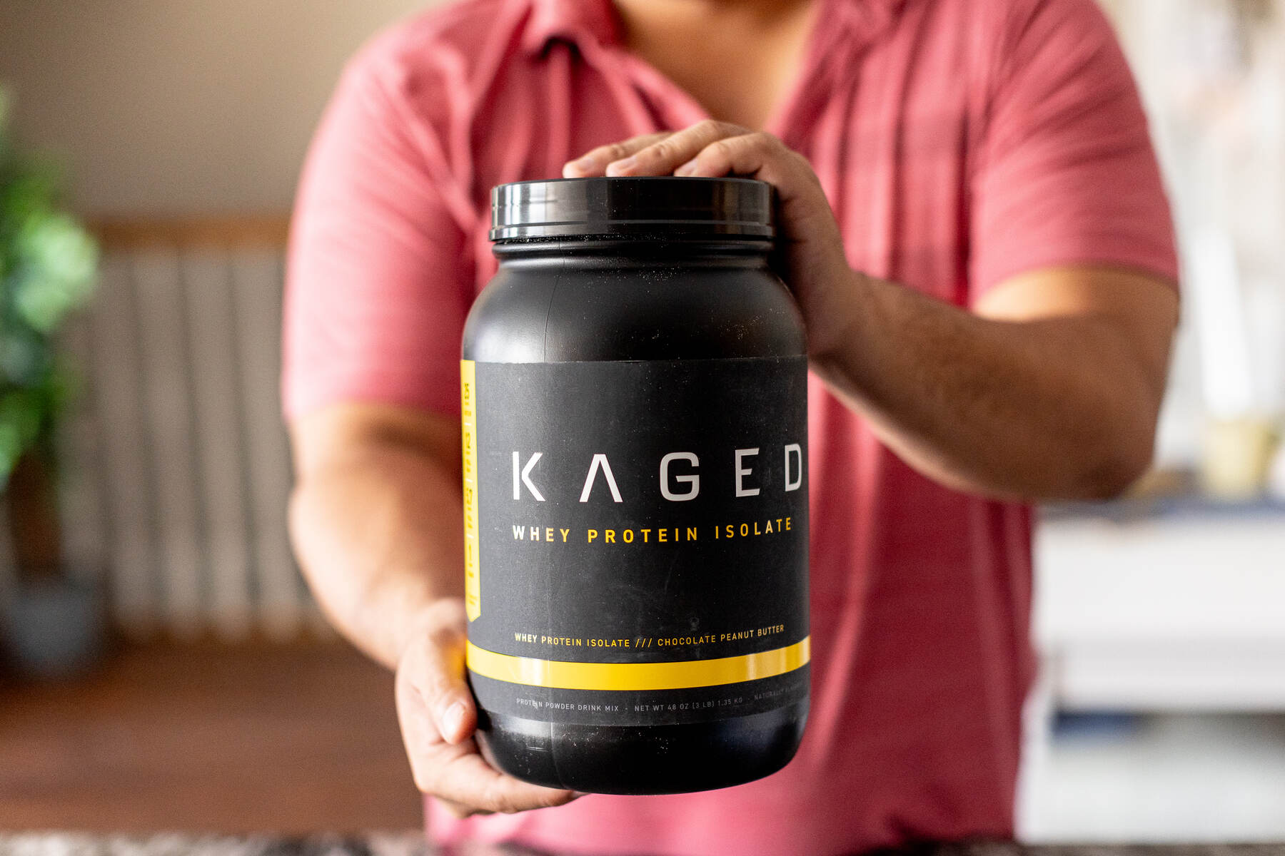 Man in a red shirt holding a large container of Kaged protein powder in a living room