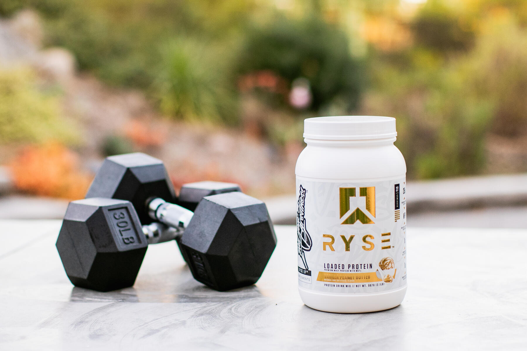 A protein powder jar with a pair of dumbbells on an outdoor marble surface